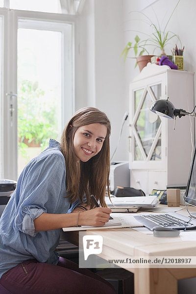 Porträt einer lächelnden jungen Frau beim Schreiben am Computertisch im Zimmer