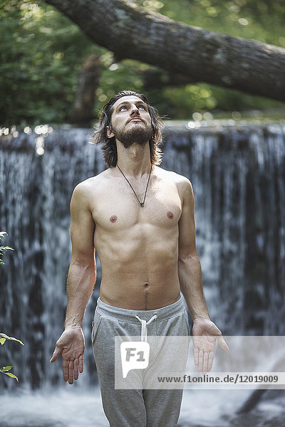Shirtless young man looking up while standing against waterfall at forest