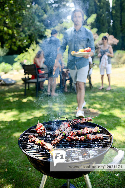 Nahaufnahme eines Grills mit Fleisch und Würstchen  die während eines Sommerfestes im Garten gegrillt werden  mit Menschen im Hintergrund.
