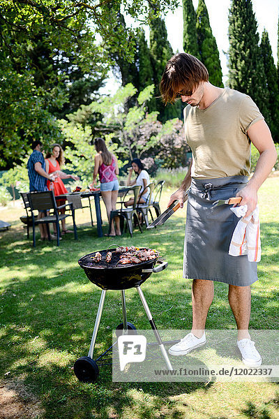 Gut aussehender junger Mann  der bei einer Grillparty im Garten während des Sommerurlaubs Fleisch zubereitet.
