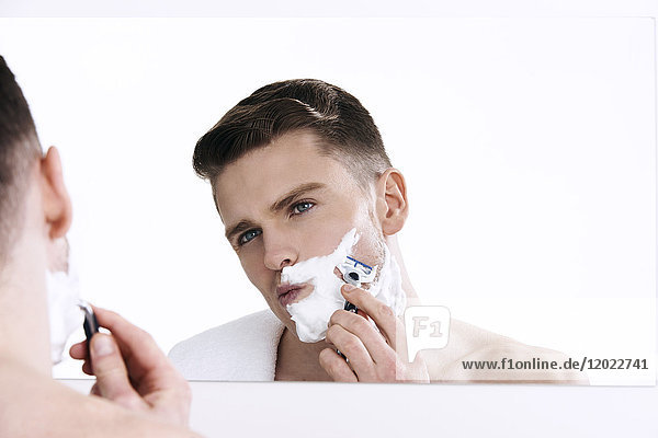 Oben-ohne-Mann  der in den Spiegel schaut  mit Rasierschaum auf seiner Gesichtshälfte  mit Rasiermesser in den Händen  rasierend