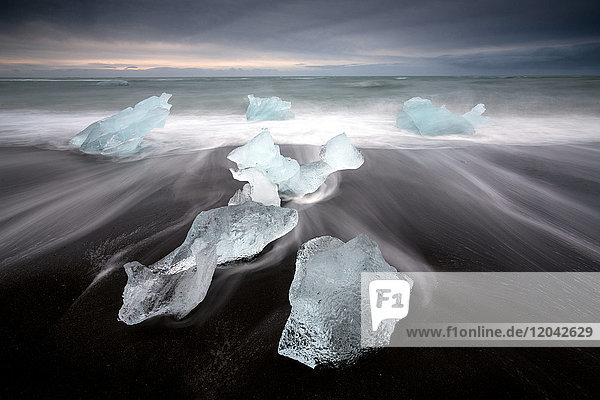 Gläserne Eisstücke auf vulkanischem schwarzem Sandstrand mit verschwommenen Wellen  in der Nähe der Lagune Jokulsarlon  Südisland  Polarregionen