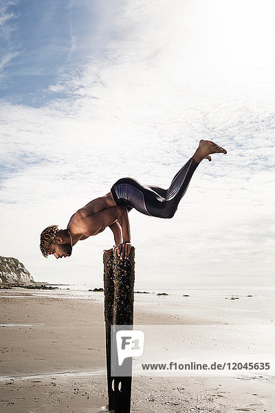 Junger Mann trainiert  macht Handstand mit erhobenen Beinen auf hölzernen Strandpfosten
