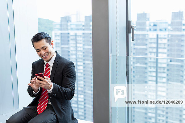 Geschäftsmann sitzt auf Fensterbank mit Smartphone und lächelt