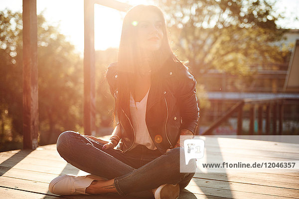 Junge Frau sitzt im Schneidersitz auf einer Holzterrasse  schaut in die Richtung  lächelt