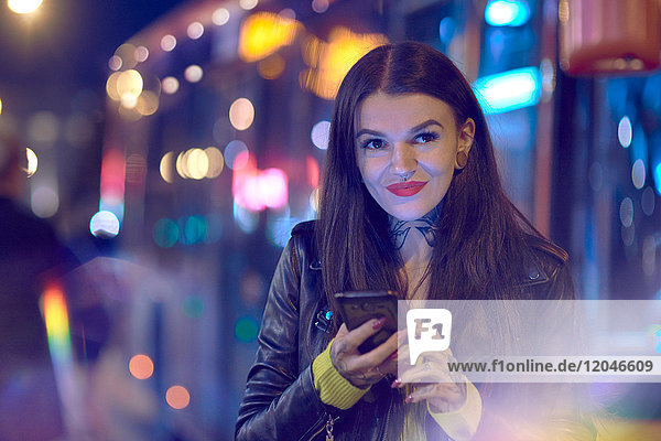 Junge Frau nachts im Freien Smartphone in der Hand haltend Tätowierungen an Hand und Hals