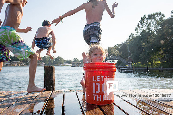 Junges Mädchen versteckt sich hinter Eimer auf Steg  Kinder springen in See