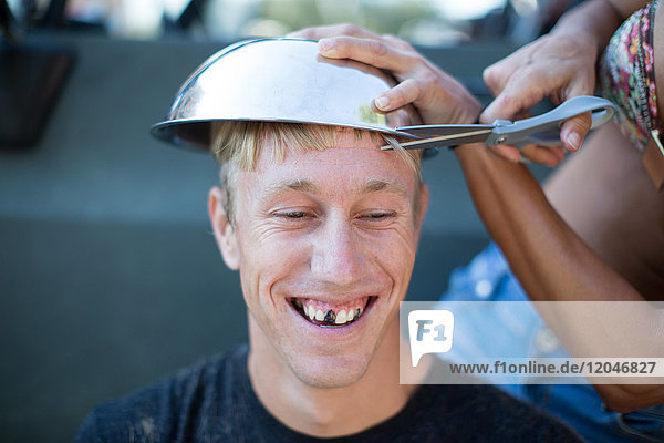 Frau schneidet jungen Männern mit Schüssel die Haare  Männern werden die Zähne geschwärzt