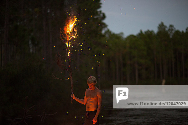 Junger Mann steht am Wasser  in der Dämmerung  hält brennenden Ast