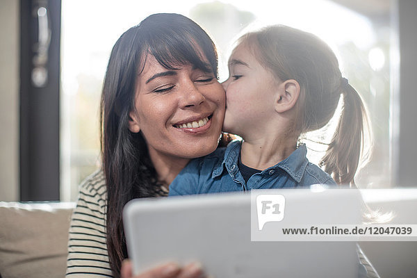 Frau wird von Tochter auf dem Sofa auf die Wange geküsst  während sie ein digitales Tablet benutzt