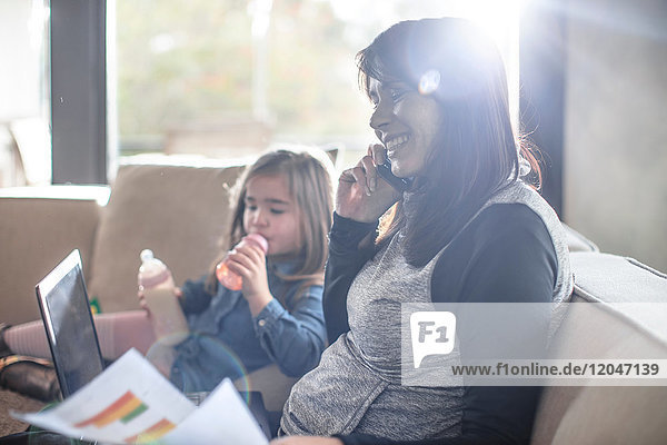 Mädchen mit Babyflasche  während die Mutter auf dem Sofa arbeitet und mit dem Smartphone telefoniert