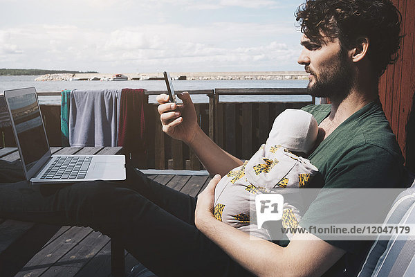 Vater mit Laptop hält Babyboy während der Benutzung des Mobiltelefons in der Ferienvilla