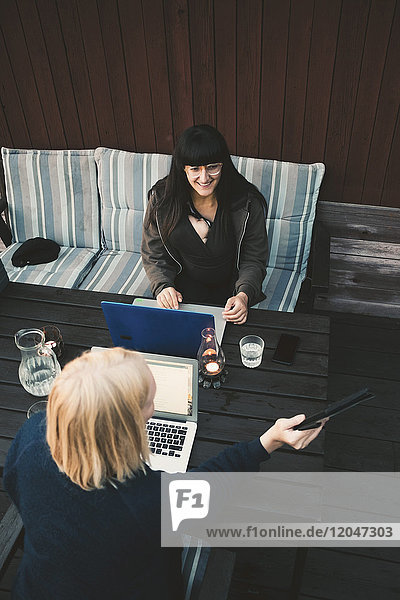 High angle view of woman showing digital tablet to female friend using laptop at table