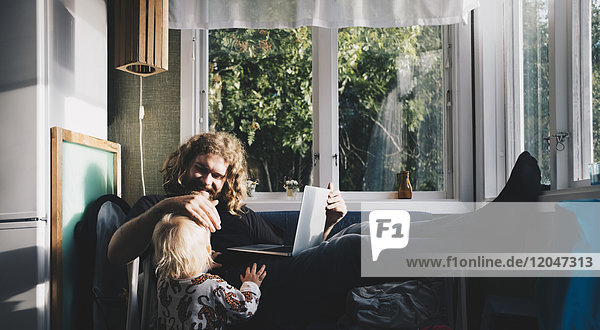 Father holding laptop while looking at girl by window