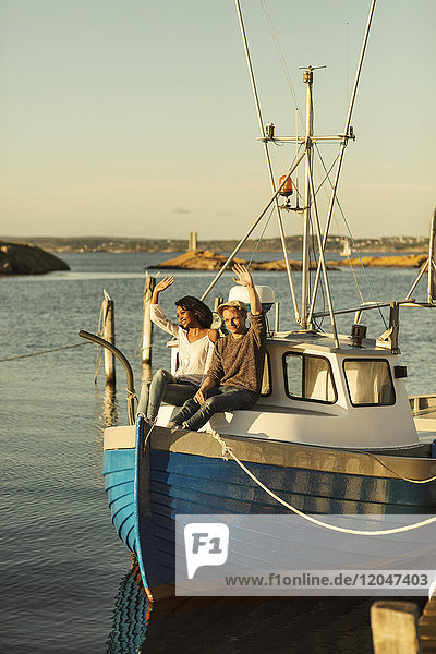 Paar mit erhobenen Händen  das im Sommer auf einem festgemachten Boot gegen den klaren Himmel sitzt.