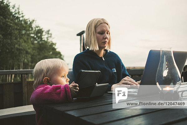Tochter hält digitales Tablett  während die Mutter den Laptop am Tisch gegen den Himmel benutzt.