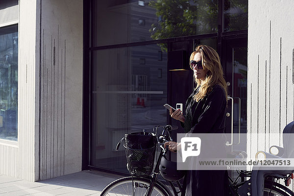 Reife Frau steht mit Handy und Fahrrad auf dem Bürgersteig gegen Gebäude in der Stadt