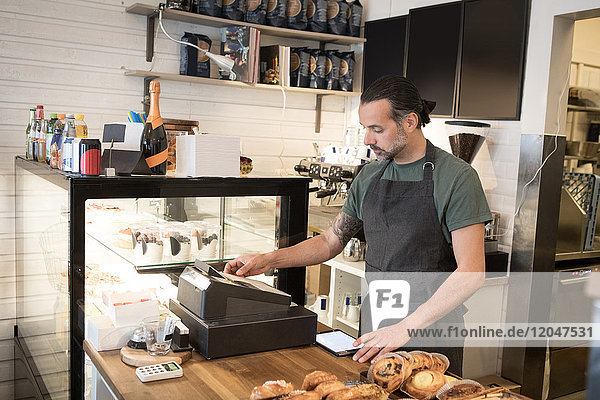 Confident mature male owner using cash register at checkout in bakery