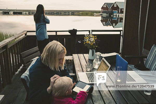 Frau unterrichtet Tochter bei der Benutzung des digitalen Tabletts  während ein Freund auf dem Handy spricht.