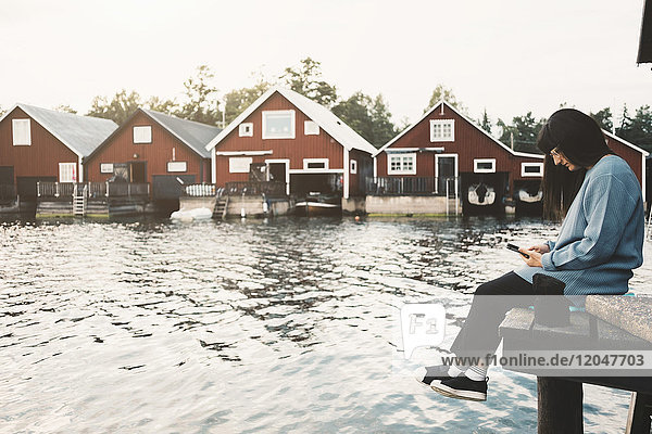 Volle Länge der Frau  die das Smartphone benutzt  während sie auf dem Steg über dem See gegen Häuser sitzt