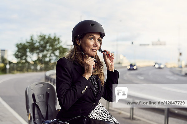 Reife Geschäftsfrau befestigt Helm auf der Straße gegen den Himmel