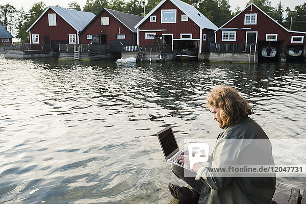 Man using laptop while sitting on jetty by lake against holiday villas