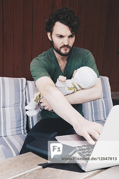 Vater mit Baby mit Laptop im Sitzen auf dem Stuhl an der Wand