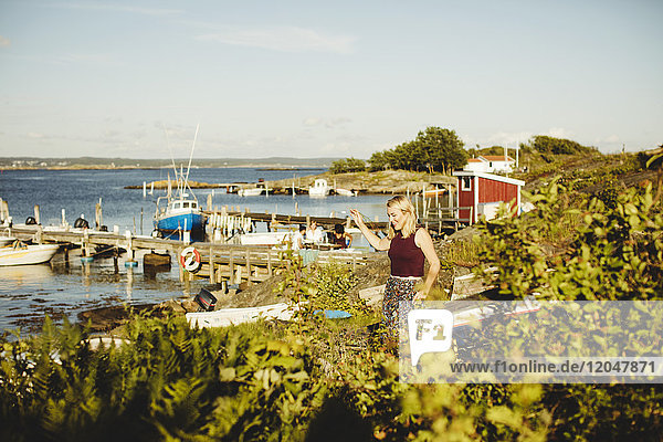 Smiling young woman walking by plants at harbor against sky on sunny day