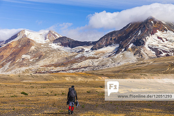 A backpacker crosses the exotic landscape of the ash and pumice-covered Valley of Ten Thousand Smokes in Katmai National Park  with Mt. Katmai (left)  Trident Volcano (right)  and the Knife Creek Glaciers looming in the distance; Alaska  United States of America