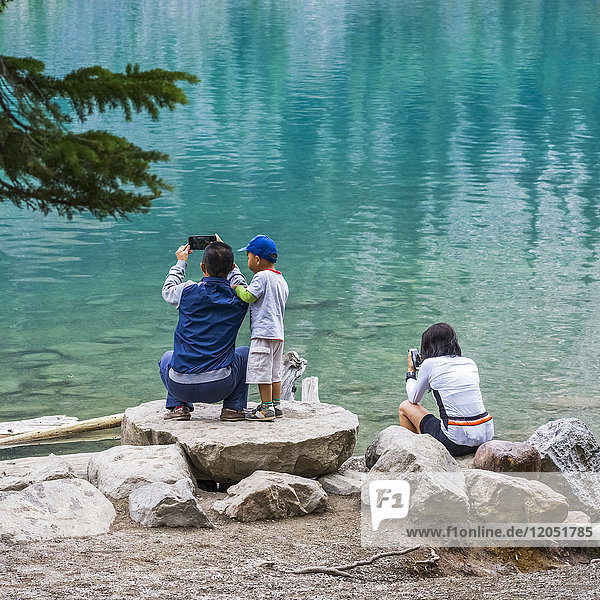 Eine Familie steht auf Felsen am Ufer des Moraine Lake und fotografiert das ruhige  türkisfarbene Wasser; Field  Alberta  Kanada
