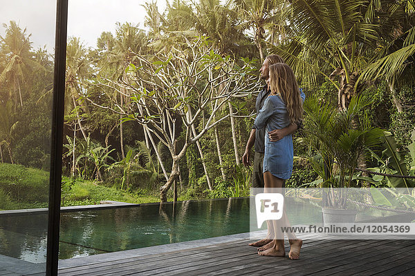Umarmendes Paar  das auf einer Holzterrasse vor dem Pool steht und eine atemberaubende Aussicht in einem üppigen tropischen Garten genießt.