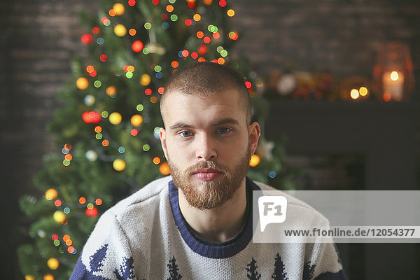 Porträt eines ernsthaften jungen Mannes mit Vollbart vor dem beleuchteten Weihnachtsbaum zu Hause