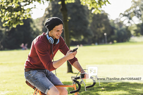 Lachender Mann auf dem Rennrad mit Blick auf das Handy im Park