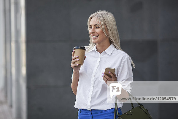 Happy businesswoman with takeaway coffee and cell phone outdoors