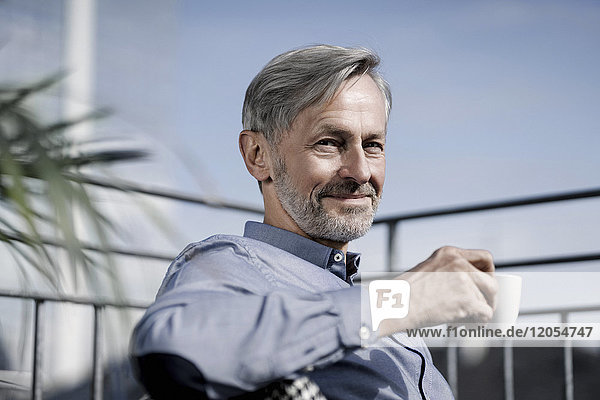 Porträt eines lächelnden grauhaarigen Mannes  der auf dem Balkon sitzt und Kaffee trinkt.