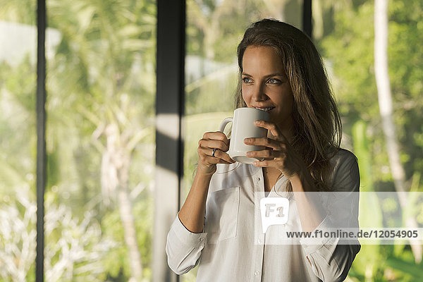 Lächelnde Frau trinkt Kaffee zu Hause vor dem üppigen tropischen Garten
