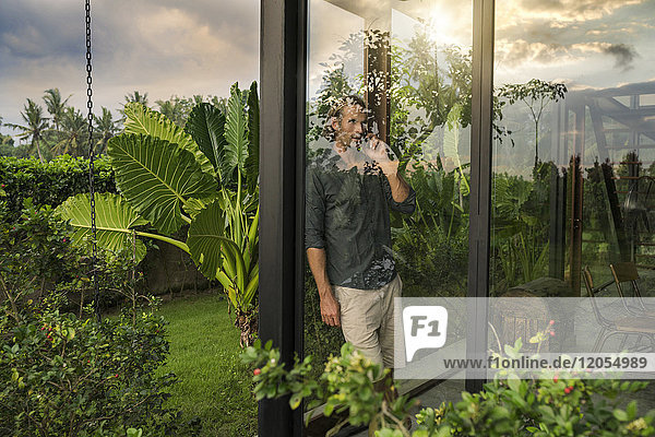 hübscher Mann  der hinter der Glasfassade des Designhauses steht und auf dem Smartphone spricht  umgeben von einem üppigen tropischen Garten.