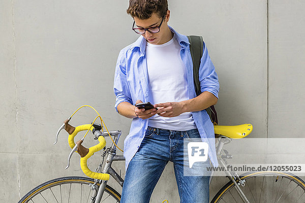 Young man with racing cycle looking at cell phone in front of concrete wall