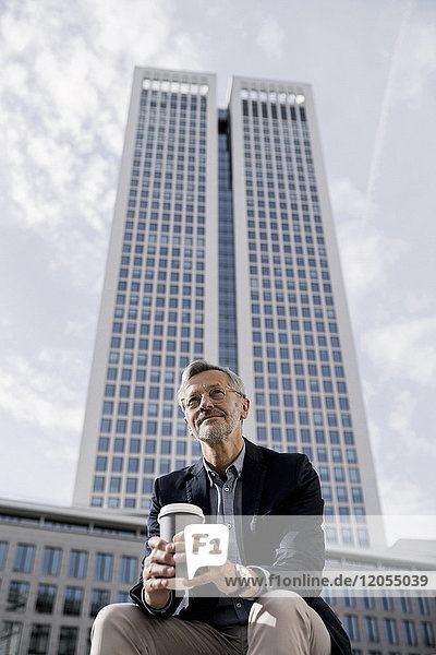Grauhaariger Geschäftsmann vor Wolkenkratzer mit Kaffee zum Mitnehmen