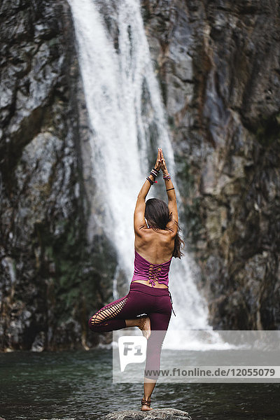 Italien,  Lecco,  Frau beim Tree Yoga Pose auf einem Felsen in der Nähe eines Wasserfalls