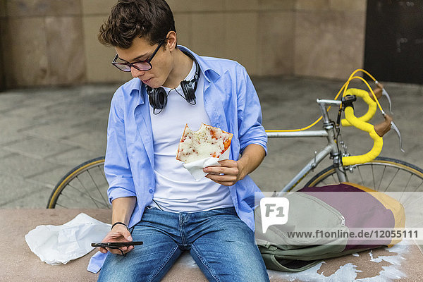 Young man eating pizza on a bench while looking at cell phone