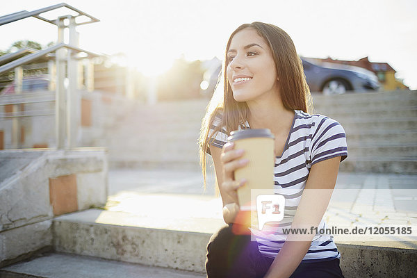 Junge Frau sitzt auf einer Treppe im Freien und hält Kaffee.