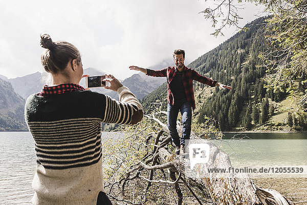 Austria  Tyrol  Alps  woman taking cell phone picture of man balancing on tree trunk at mountain lake