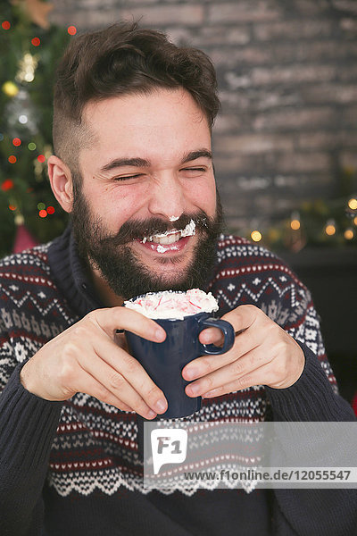 Porträt eines lachenden Mannes  der heiße Schokolade mit Schlagsahne und gehackten Zuckerstangen trinkt.