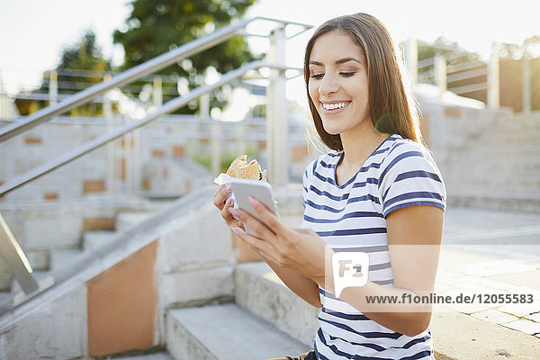 Junge Frau sitzt auf einer Treppe und isst Bagel und benutzt ein Smartphone.