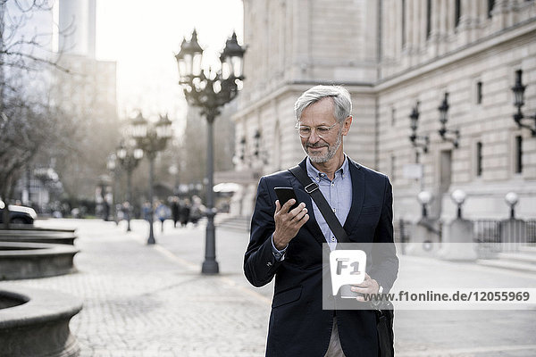 Grauhaariger Geschäftsmann in der Stadt mit Smartphone und Kaffee zum Mitnehmen