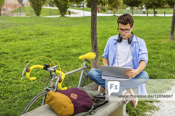 Young man with racing cycle sitting on a bench using laptop