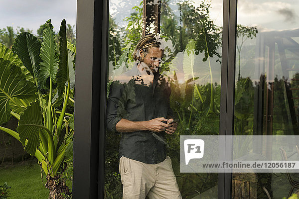 Schöner Mann steht hinter der Glasfassade des Designhauses und schaut auf das Smartphone  umgeben von einem üppigen tropischen Garten.