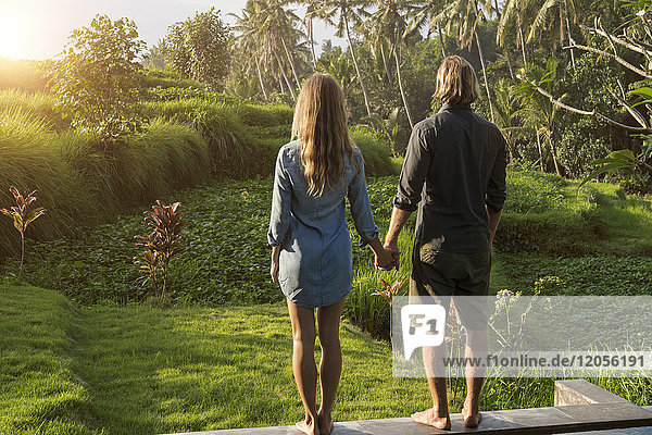 Ein Paar hält sich an den Händen  während es am Rande eines Pools steht und einen atemberaubenden Blick auf den Sonnenuntergang in einem üppigen tropischen Garten genießt.
