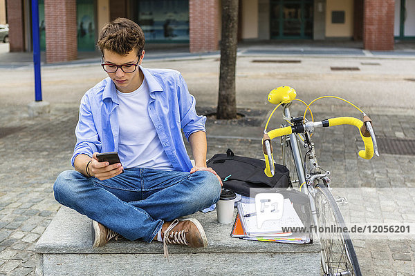 Junger Mann mit Rennrad sitzt auf der Bank und schaut aufs Handy.